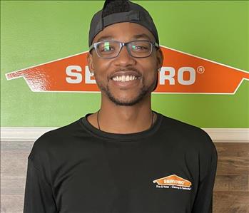 Male employee with a hat and glasses smiling in front of green SERVPRO background.
